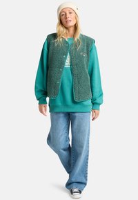 Teal fleece-vesti teal-sweatshirta päällä, yhdistettynä vaaleansinisiin leveälahkeisiin farkkuihin ja mustiin tennareihin. Västissä on napit ja teksturoitu pinta.