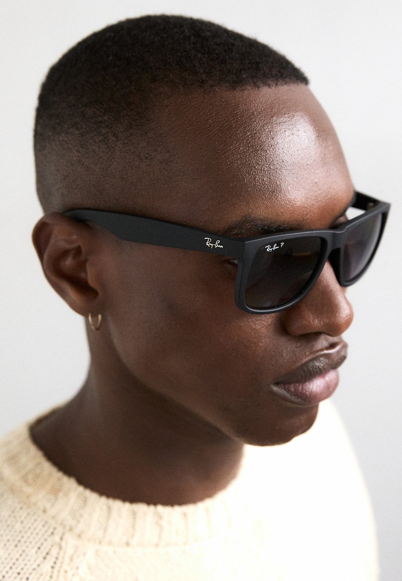 Ochelari de soare rectangulari negri cu lentile întunecate. Ramele sunt mate și au o marcă subtilă Ray-Ban. Purtați de o persoană cu păr scurt.