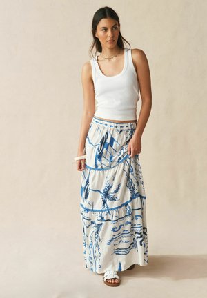 Kobieta o długich ciemnych włosach, ubrana w białą bezrękawnik, niebiesko-białą spódnicę maxi w wzory, białe sandały i bransoletkę, stojąca na beżowym tle.