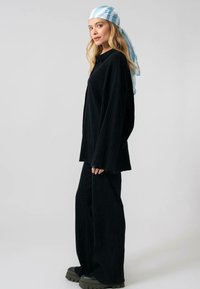 Juodas raukšlėtas loungewear komplektas su laisvu kirpimu. Marškiniai turi ilgas rankoves ir mygtuką, derinami su plačiomis kelnių kelnėmis ir masyviais juodais batais.