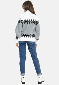 Kobieta zwr�ócona plecami, ubraną w biało-szary sweter we wzory, niebieskie dżinsy z podwiniętymi nogawkami oraz białe botki z czarną podeszwą.