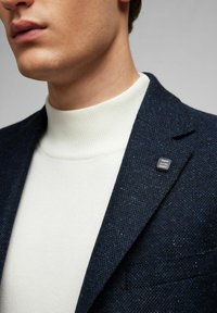 Navyfarbener, strukturierter Blazer mit einem weißen, gerippten Rollkragenpullover darunter; verfügt über eine kleine schwarze Markennadel am Revers.