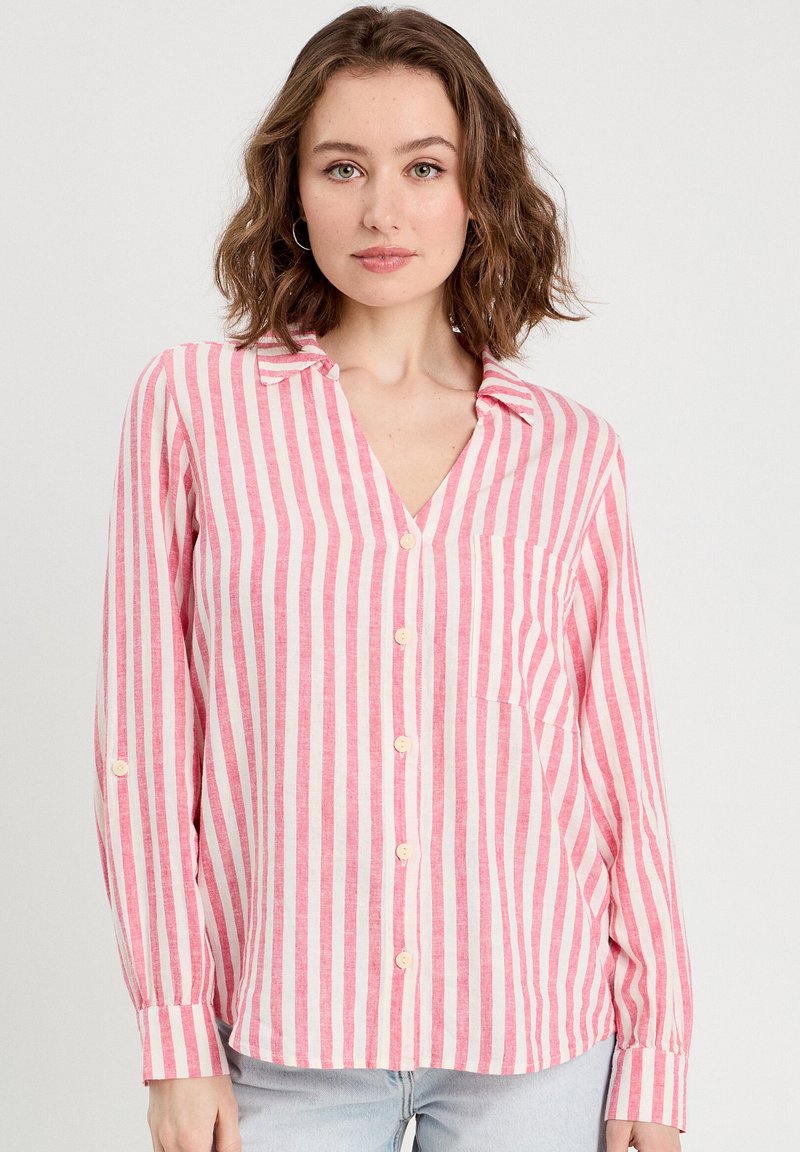 Chemise à manches longues rayée rose et blanche avec un col, des boutons frontaux discrets et une poche sur la poitrine. Texture de tissu douce.