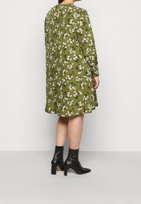 Abito floreale verde con maniche lunghe, caratterizzato da un dettaglio a oblò nella schiena e una forma svasata. Indossato con stivaletti neri alla caviglia.