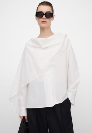 Weißes Baumwollhemd mit asymmetrischem, drapiertem Design, Knopfverschluss und weiten Ärmeln, kombiniert mit schwarzer Hose und übergroßen Sonnenbrillen.