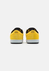 Căști de skateboard din suede galben, cu o căptușeală interioară neagră, talpă intermediară albă și talpă exterioară neagră. Logo-ul "etnies" brodat pe partea din spate.