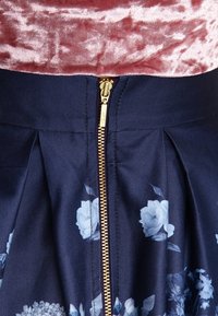 Laivastonsininen kukkamekko, jossa on sinisiä ruusukuoseja, muotoiltuja poimuja ja kultainen vetoketjulukko vyötäröllä. Silkkinen kangas.