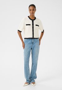 Short-sleeve, cream cardigan with black trim and pockets, paired with light blue jeans and off-white pointed shoes. Smooth texture, casual fit.