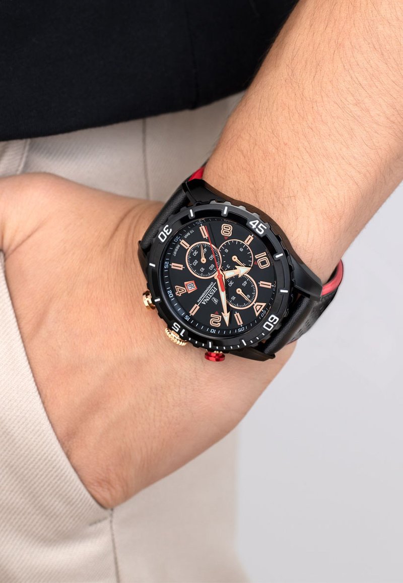 Montre analogique noire avec des cadrans multifonctionnels, des accents rouges, des boutons en or et en argent, et un bracelet en cuir noir, portée au poignet.
