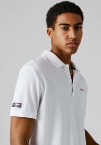 Νέος άνδρας που φοράει λευκό πόλο μπλουζάκι Red Bull Racing, στραμμένος ελαφρώς προς τα δεξιά με ουδέτερη έκφραση, σε απλό ανοιχτό γκρι φόντο.