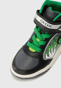 Sneaker neri e grigi con finitura texturizzata, lacci verdi, branding argentato e suola bianca con grafiche accentuate.