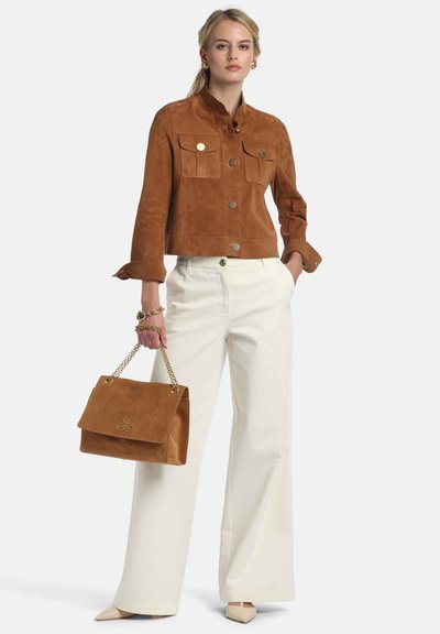 Braune Wildlederjacke in Cropped-Optik mit goldfarbenen Knöpfen und Taschen, kombiniert mit weißen, weit geschnittenen Hosen und einer passenden braunen Handtasche mit Kettenriemen.