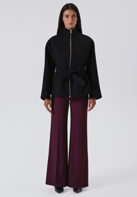 Giacca nera con zip e cintura, caratterizzata da maniche ampie e due tasche anteriori; abbinata a pantaloni bordeaux a vita alta e gamba larga.