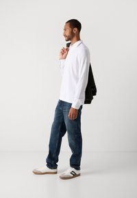 Mann in weißem Langarm-Polohemd, blauen Jeans und weißen Turnschuhen, der eine dunkle Jacke über der Schulter hält und vor einem schlichten hellen Hintergrund steht.