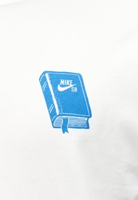 Μπλε απεικόνιση ενός κλειστού βιβλίου με σελιδοδείκτη και το κείμενο "NIKE SB" μαζί με το λογότυπο swoosh σε λευκό υφασμάτινο φόντο.