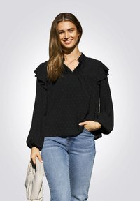 Zwarte blouse met lange mouwen, versierd met ruches, gestructureerde stippen en een V-halslijn, gecombineerd met blauwe jeans en een lichtgekleurde clutch in de hand.
