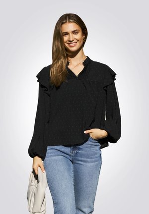 Zwarte blouse met lange mouwen, versierd met ruches, gestructureerde stippen en een V-halslijn, gecombineerd met blauwe jeans en een lichtgekleurde clutch in de hand.