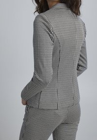 Räätälöity bleiseri mustavalkoisella koirankynsimuodolla. Siinä on rakenteelliset hartiat, istuva siluetti ja sileä kangaspinta.