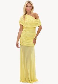 Femme portant une longue robe jaune asymétrique froncée en tissu transparent, avec un bracelet et une bague en or, debout devant un fond uni.