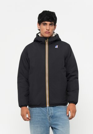 Veste rembourrée noire avec une fermeture éclair jaune et blanche, capuche avec cordon de serrage et logo sur la poitrine. Portée avec un jean bleu clair.
