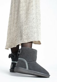 Botas de tobillo de ante gris con punta redonda, con un acento metálico, detalle de lazo en la parte trasera y suela texturizada. Se llevan con una falda estampada.