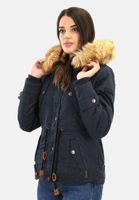 Vrouw die de bontomzoomde capuchon van een marineblauwe winterjas met knopen en ritsdetails aanpast, terwijl ze een spijkerbroek draagt tegen een effen achtergrond.