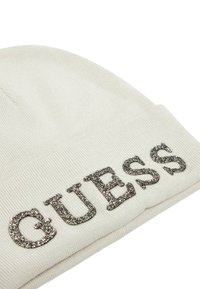 Berretto lavorato a maglia bianco con lettering "GUESS" in rilievo e glitterato. Texture morbida con una vestibilità aderente, ideale per il freddo.