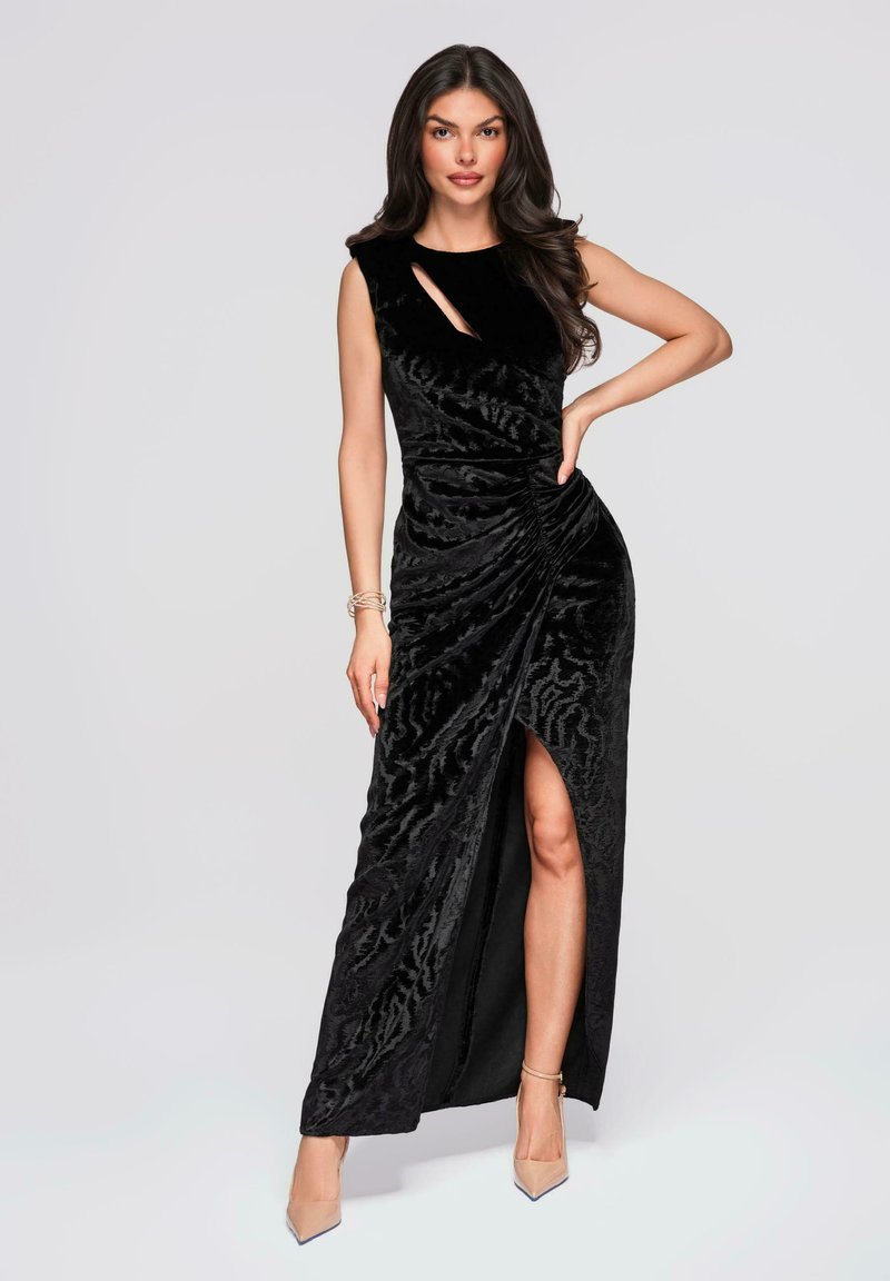 Femme posant dans une robe sans manches en velours noir avec une fente haute froncée et un décolleté en trou de serrure, portant des escarpins nude pointus et des bracelets.