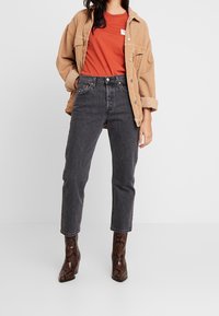 Brun kordflodenjacka över en orange t-shirt, tillsammans med svarta rak benjeans och bruna ankelboots med ormskinnsmönster.