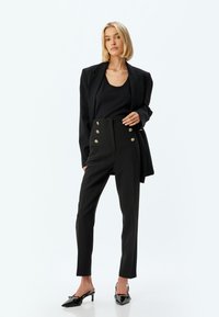 Blazer nero su un top nero, pantaloni neri a vita alta con dettagli nei bottoni dorati e scarpe con tacco nere.