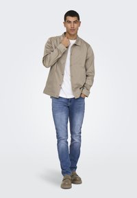 Ελαφρώς μπεζ σακάκι με κουμπιά πάνω από λευκό t-shirt, συνδυασμένο με μπλε τζιν και καφέ παπούτσια, στέκεται μπροστά σε ουδέτερο φόντο.