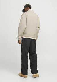 Sudadera beige con cuello alto y corte oversized, combinada con pantalones cargo negros con múltiples bolsillos y calzado marrón.