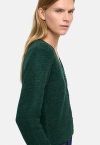 Zielony sweter z dzianiny, z dekoltem w serek, guzikami z przodu i długimi rękawami. Materiał wydaje się miękki, o subtelnej teksturze i ciepłym odcieniu.