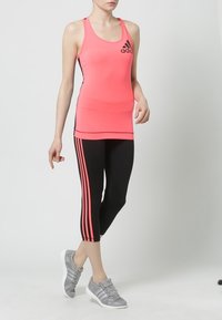 Woman wearing a coral Adidas tank top, black cropped leggings with coral stripes, and gray Adidas sneakers, standing against a plain background.