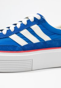 Blue canvas sneaker with white stripes, suede accents, white laces, and a thick white sole featuring a red stripe along the bottom edge.