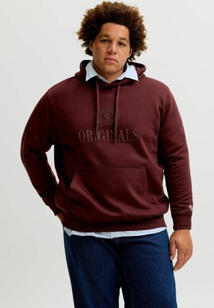 Mann mit lockigem Haar und Sommersprossen, der einen dunkelroten Kapuzenpullover mit der Aufschrift „ORIGINALS“ und dunkelblaue Jeans vor einem schlichten Hintergrund trägt.