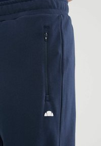 Zbliżenie granatowych spodni dresowych z pionową kieszenią na zamek błyskawiczny oraz małym białym logo w kształcie półokręgu w pobliżu kieszeni po prawej stronie.