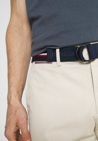 Meripihan navy dengan gesper logam hitam bulat, dihiasi dengan aksen garis merah dan putih, dikenakan di atas celana khaki.