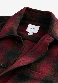 Veste en laine à carreaux rouges et noirs avec une fermeture à bouton, une texture douce et une étiquette affichant la marque "NEXT" à l'intérieur du col.