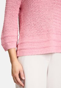 Maglione rosa lavorato a maglia con una trama testurizzata e apertura, maniche a tre quarti e orlo a coste. Abbinato a pantaloni chiari.