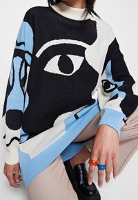 Maglione lavorato a maglia in bianco e nero con grafica di un occhio blu. Presenta un collo alto e polsini a costine, abbinato a pantaloni beige e anelli colorati.