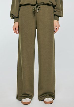 Spodnie o szerokich nogawkach w oliwkowej zieleni ze ściągaczem w pasie, noszone z białymi trampkami na stojącej osobie.