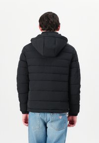 Geacă puffer neagră cu glugă, cu un design matlasat și manșete elastice, purtată peste blugi albaștri cu un eticheta pe buzunarul de la spate.