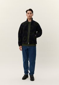 Υφασμάτινο σκούρο μπλε τζάκετ με φερμουάρ και πράσινες λεπτομέρειες, φορεμένο πάνω από πράσινο τοπ, σε συνδυασμό με μπλε τζιν και μαύρα παπούτσια.