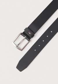 Czarny skórzany pasek z prostokątną srebrną klamrą i małym prostokątnym logo w kolorze czerwonym, białym i granatowym blisko zapięcia na białym tle.