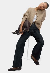 Mujer vistiendo un cárdigan de punto beige, blusa blanca, pantalones de pierna ancha oscuros y bailarinas oscuras, sosteniendo un gran bolso de piel marrón oscuro.