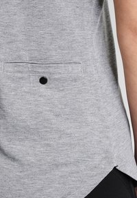 Camisa gris de manga corta con textura suave, que cuenta con un bolsillo frontal delineado con costuras y un botón negro como detalle.