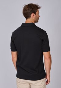 Polo noir à manches courtes, col côtelé et design épuré. Le matériau semble lisse, avec une coupe ajustée et des fentes sur les côtés au niveau de l'ourlet.