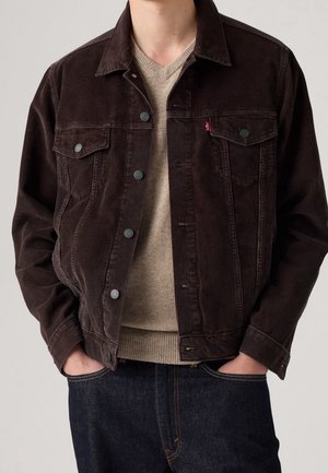 Hombre con chaqueta de pana marrón oscuro sobre un suéter beige de cuello en V, con las manos en los bolsillos de unos vaqueros azul oscuro.