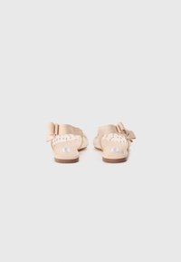 Light beige sandals with a textured upper, featuring a large bow and cut-out details on the sides, set on a flat brown sole.